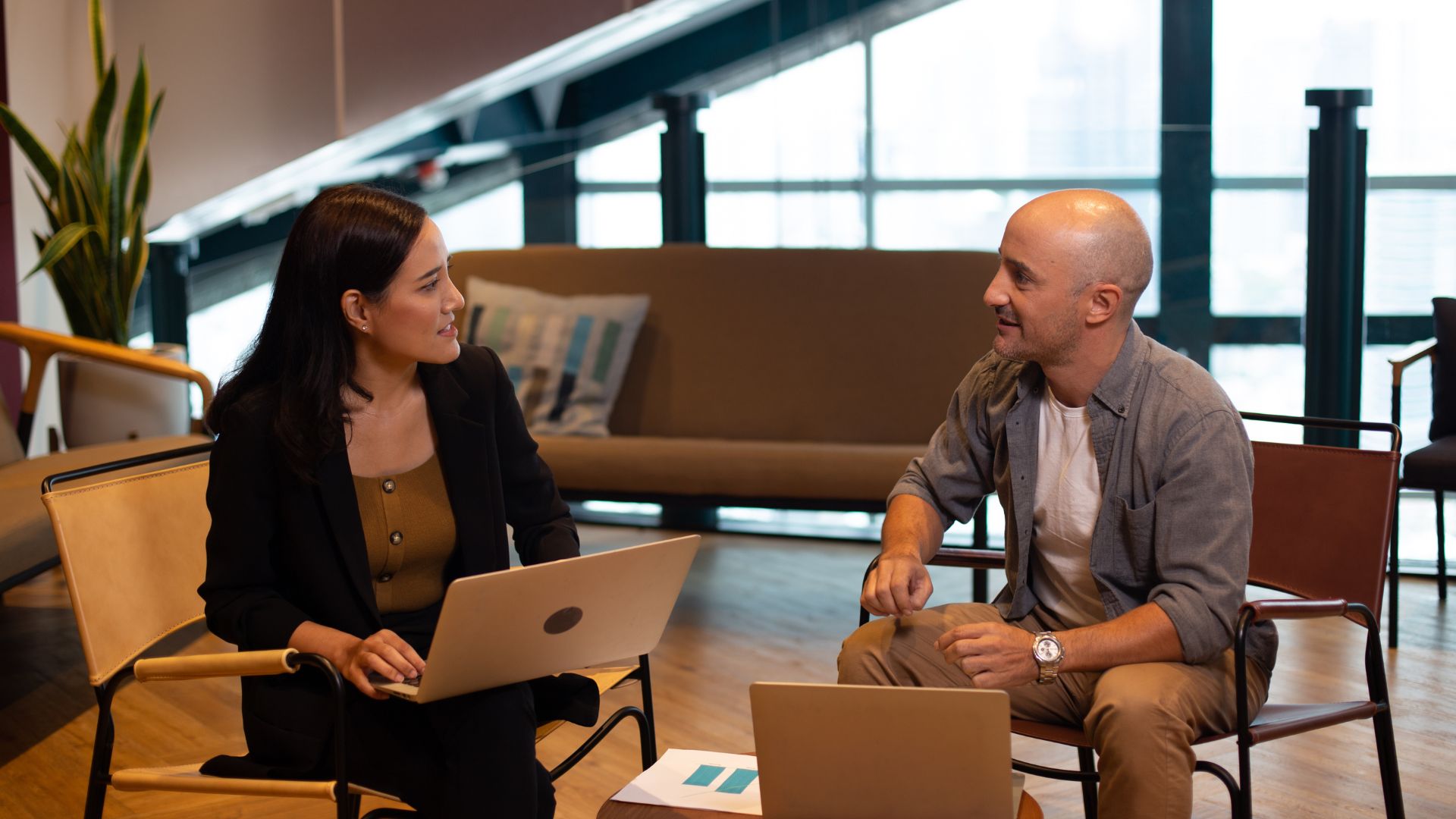 A woman and a bald man sit facing each other, each with a laptop, in a modern office setting with large windows and a couch in the background. They are engaged in a weekly one-on-one meeting, discussing work with attention and ease.