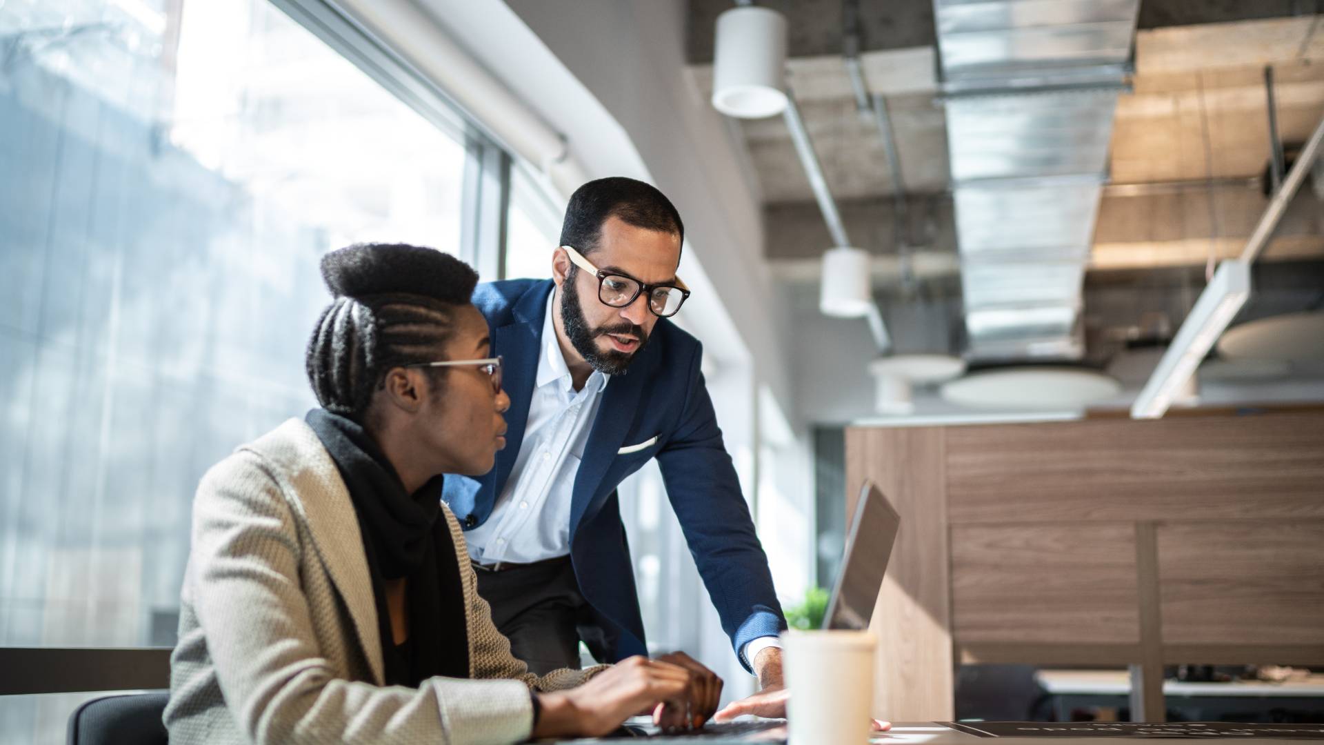 There's a woman sitting at a desk with a laptop in front of her. She's looking up at a man who is speaking to her and is standing next to her. They are both wearing glasses.