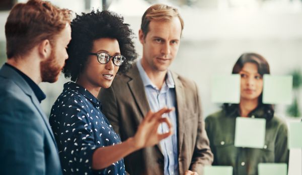 A diverse team of four people are talking in front of a transparent white board with post it notes on it.