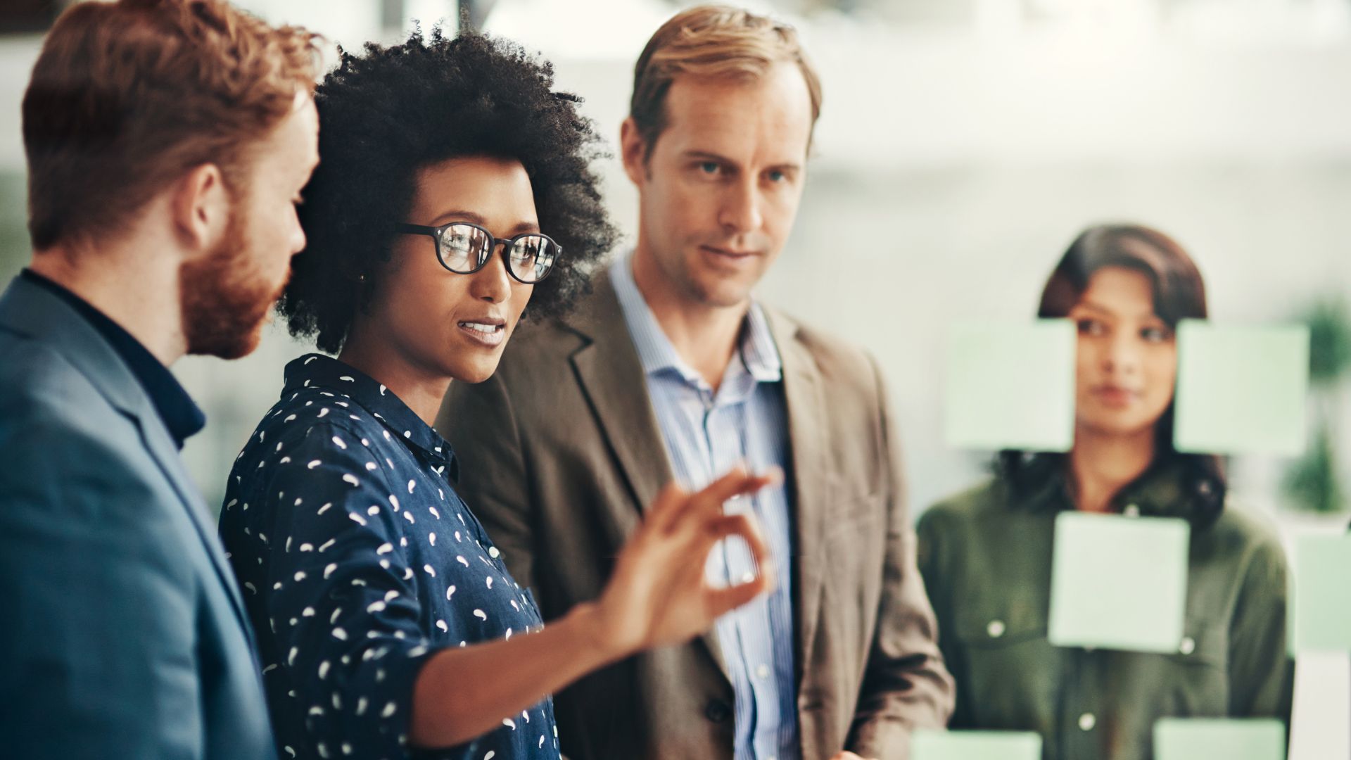 A diverse team of four people are talking in front of a transparent white board with post it notes on it.
