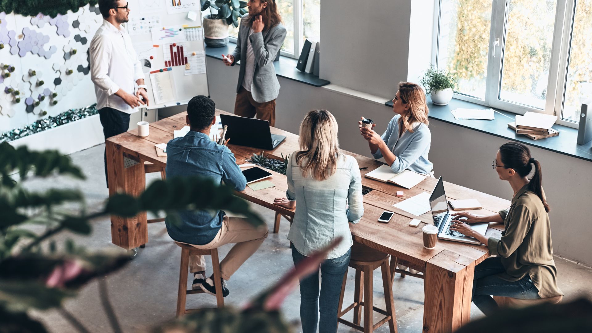 A diverse team of people are standing and sitting around a tall conference table looking at two men in front of a flip chart during a brainstorming session.