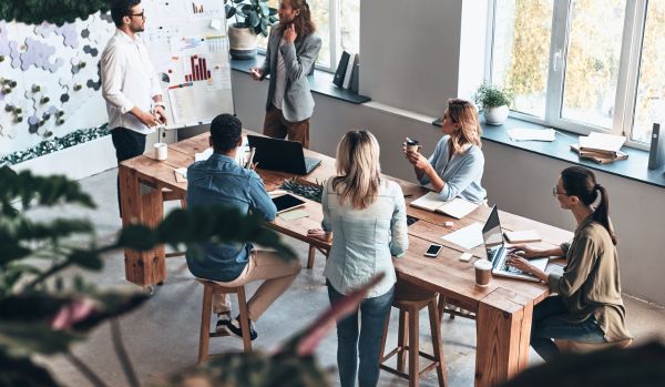 A diverse team of people are standing and sitting around a tall conference table looking at two men in front of a flip chart during a brainstorming session.