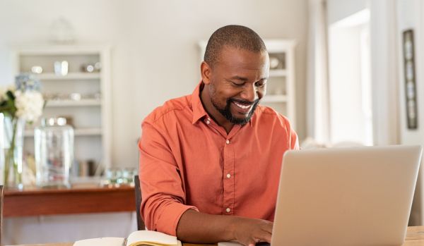 A man is sitting at his dining room table working on his laptop and smiling.