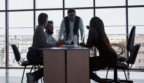 A diverse team is gathered around a table discussing a project. The person leading the conversation is standing and implementing skills learned in custom simulations.