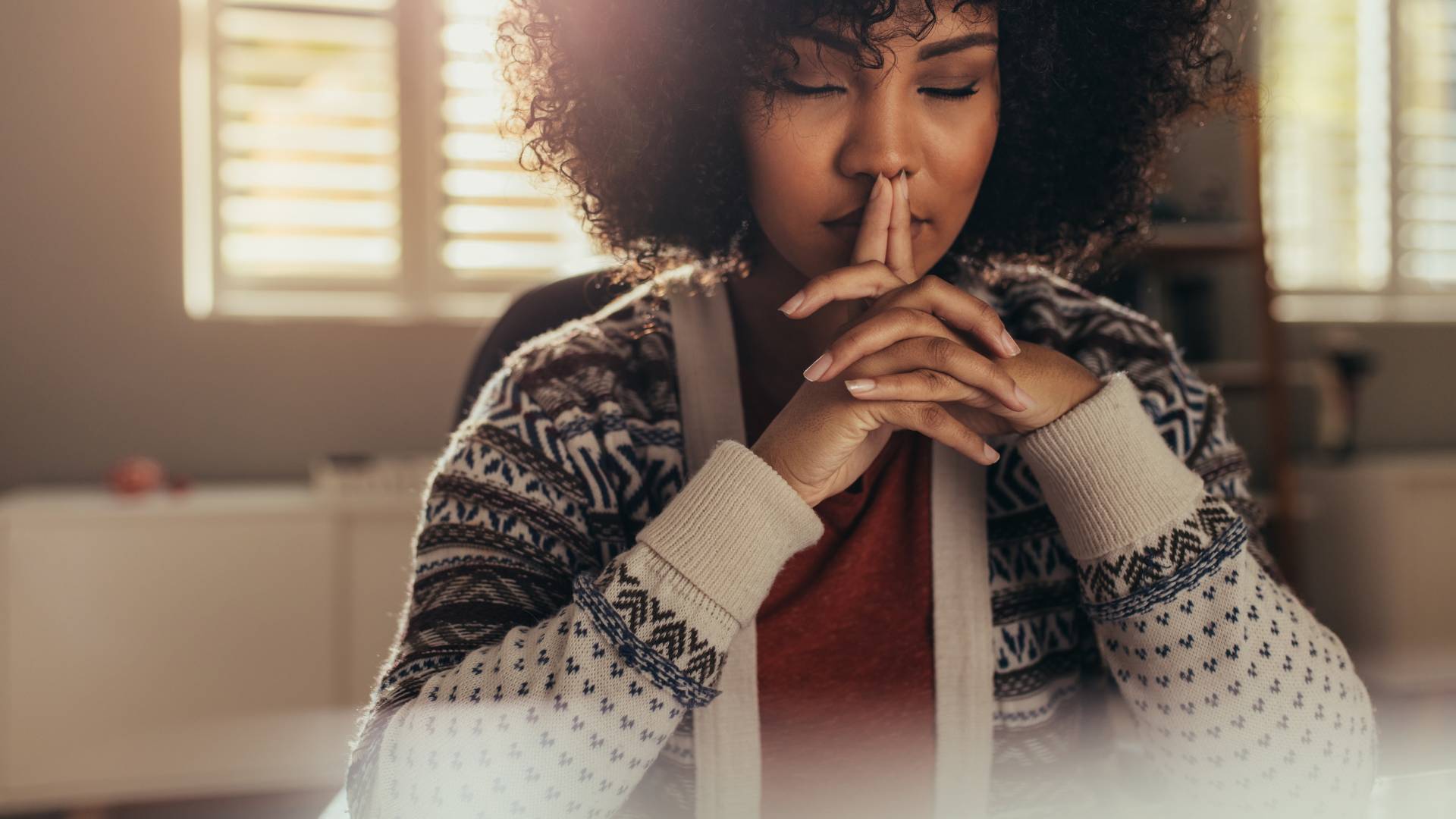 A woman sits with her elbows on the table, hands clasped together. Her eyes are closed as she rests her chin on her hands.