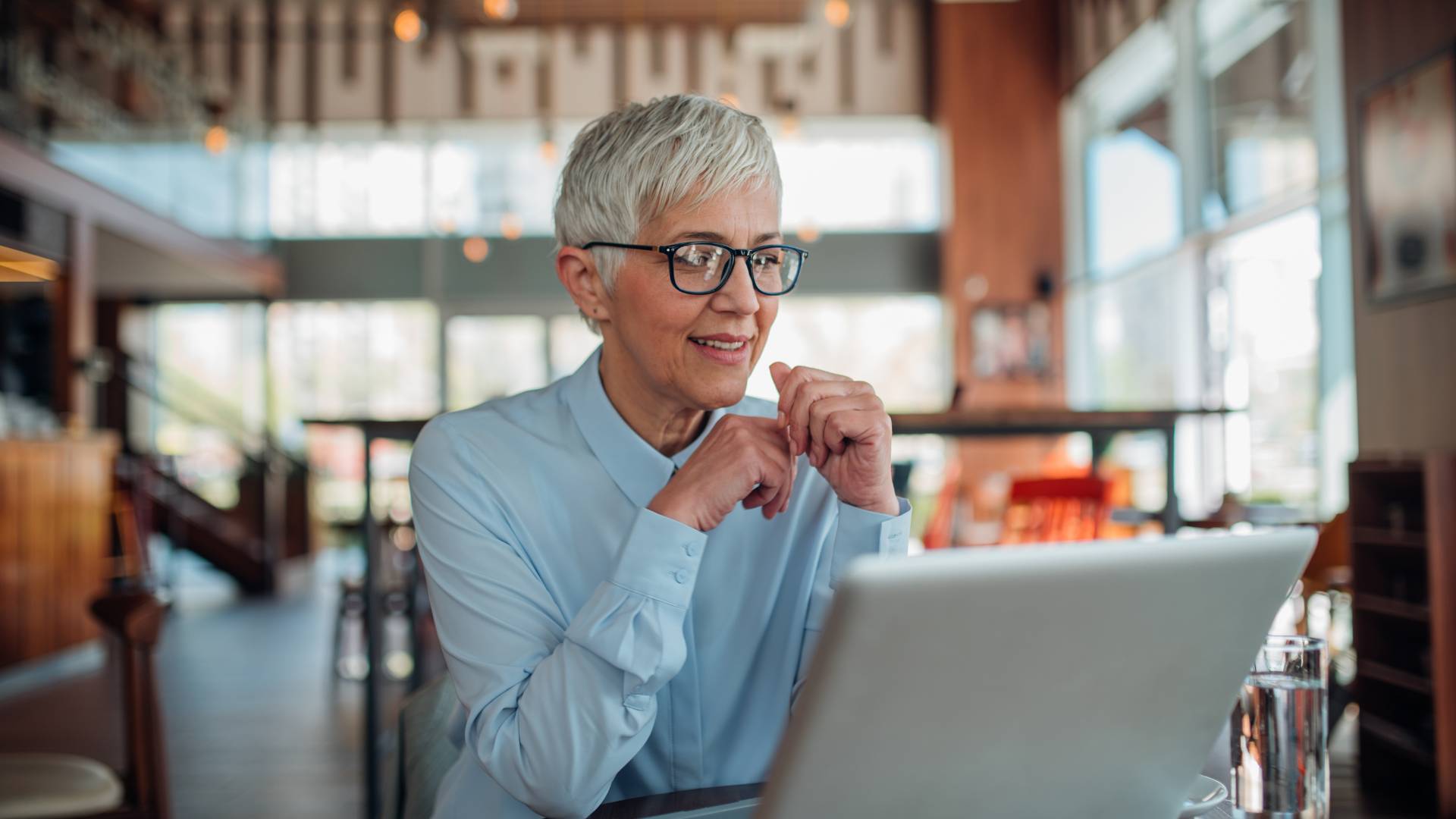 An older woman with a short, gray pixie cut is working on her laptop in an office common space.