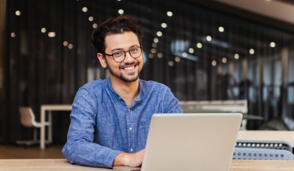 A happy young office worker is smiling at the camera over his laptop in an open working space.