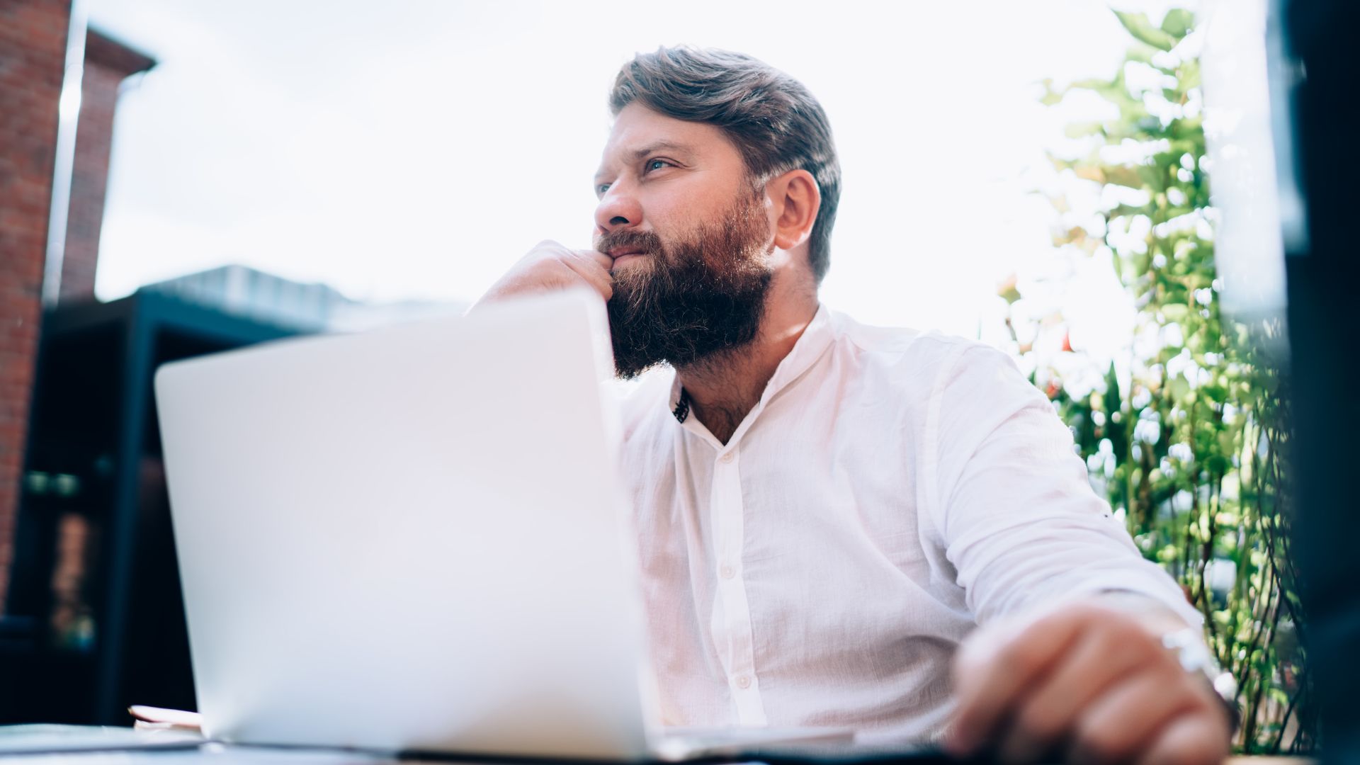 A bearded man is sitting outside on his laptop with a pensive look on his face.
