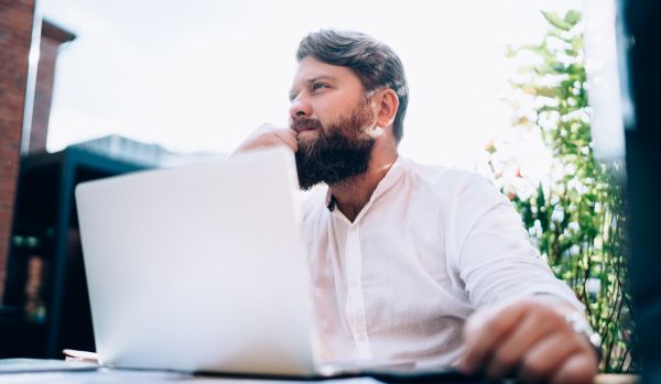 A bearded man is sitting outside on his laptop with a pensive look on his face.