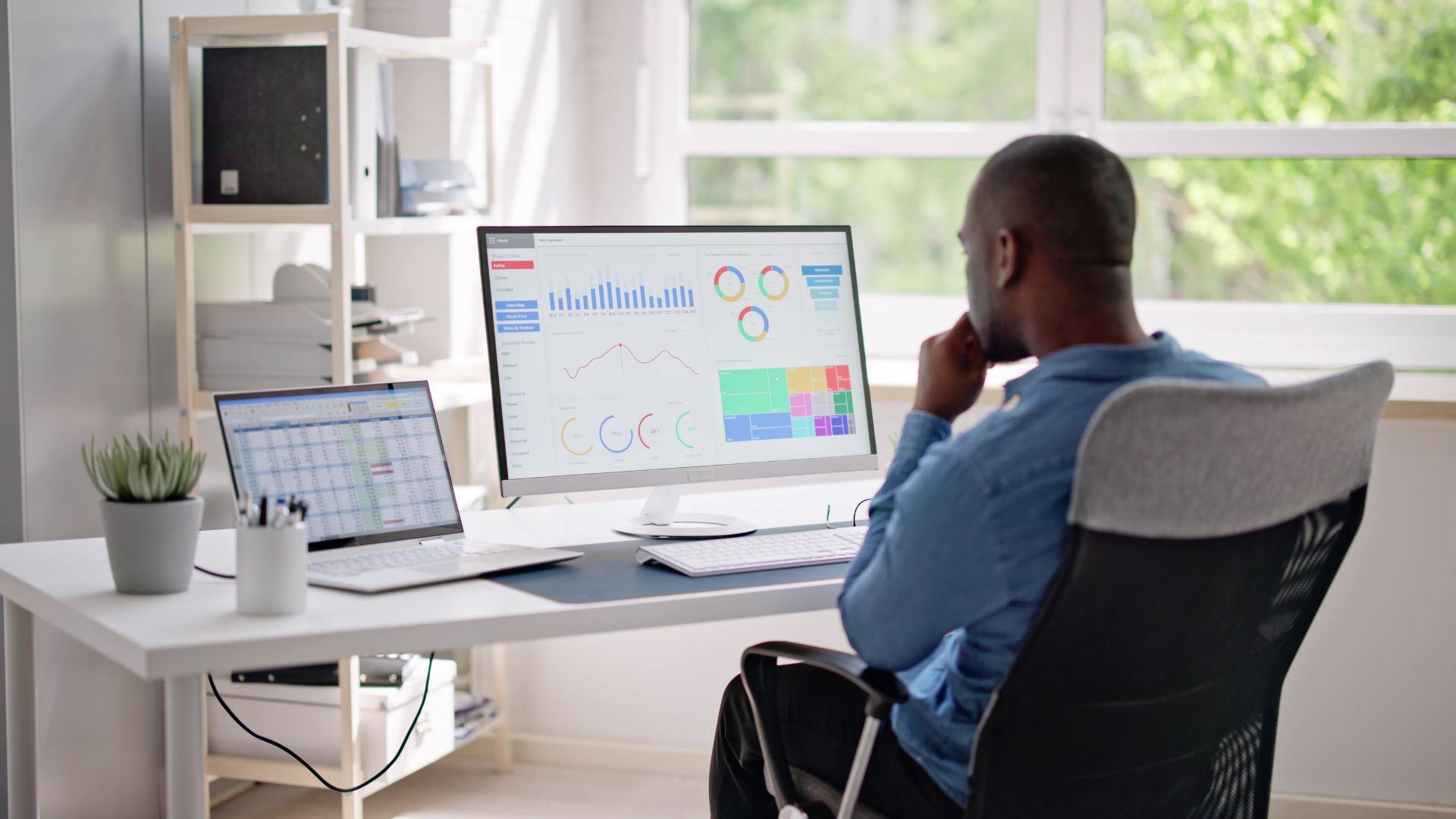 We're looking over the shoulder of a man reviewing a data dashboard on his monitor in his home office.