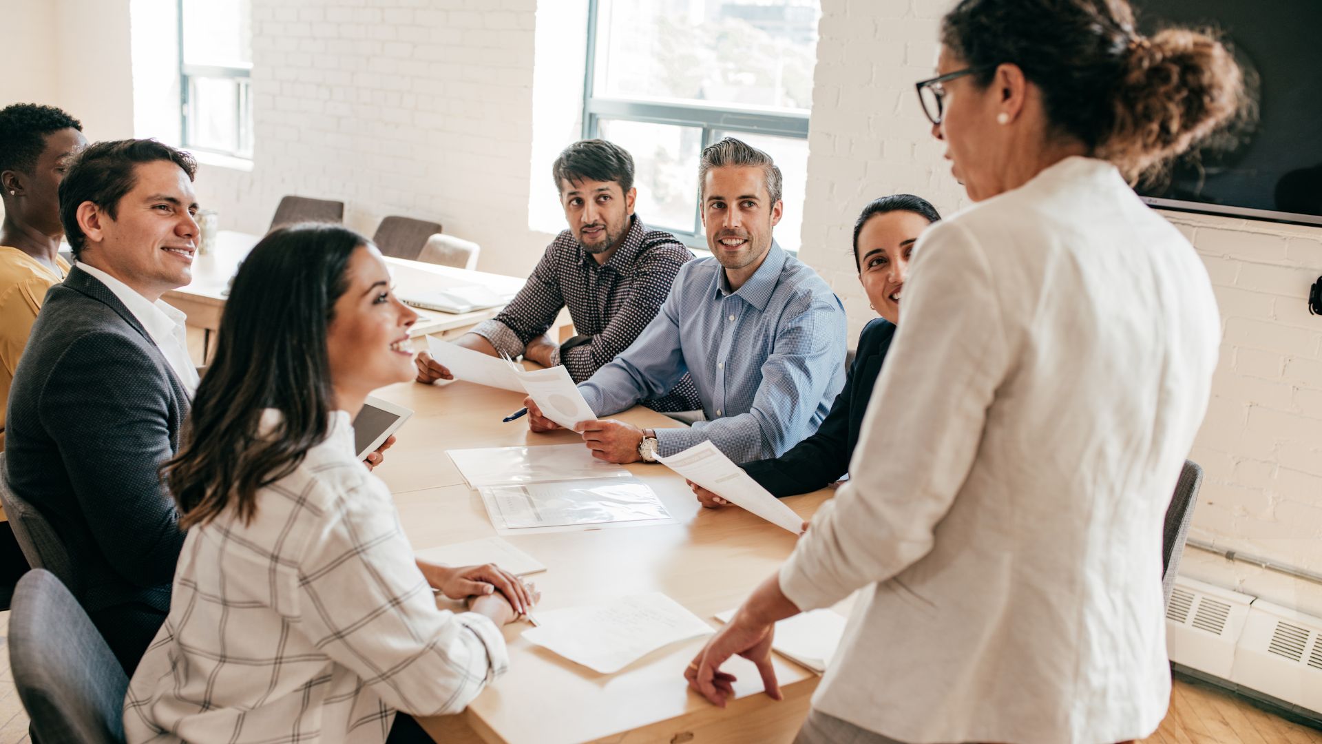 A woman is standing at the front of a conference table where diverse aspiring leaders are listening to her present.