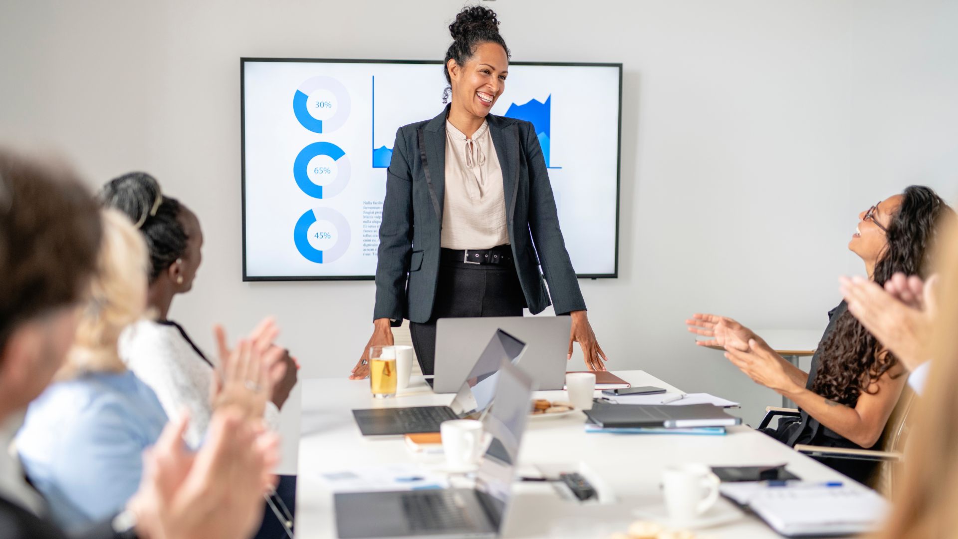 A team is seated around a conference table clapping for the woman standing at the end who just finished a presentation.