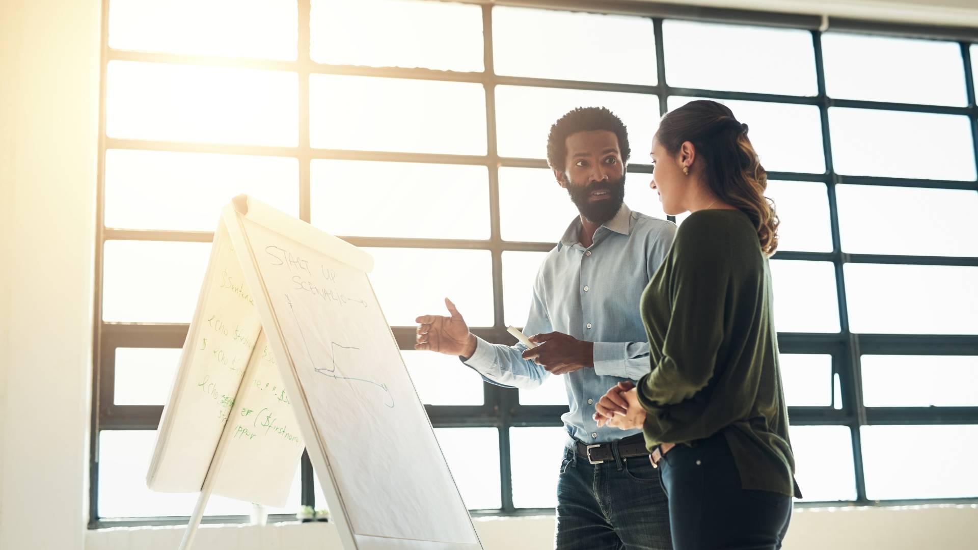 A mand and a woman are standing in front of a flipchart doing some scenario planning.