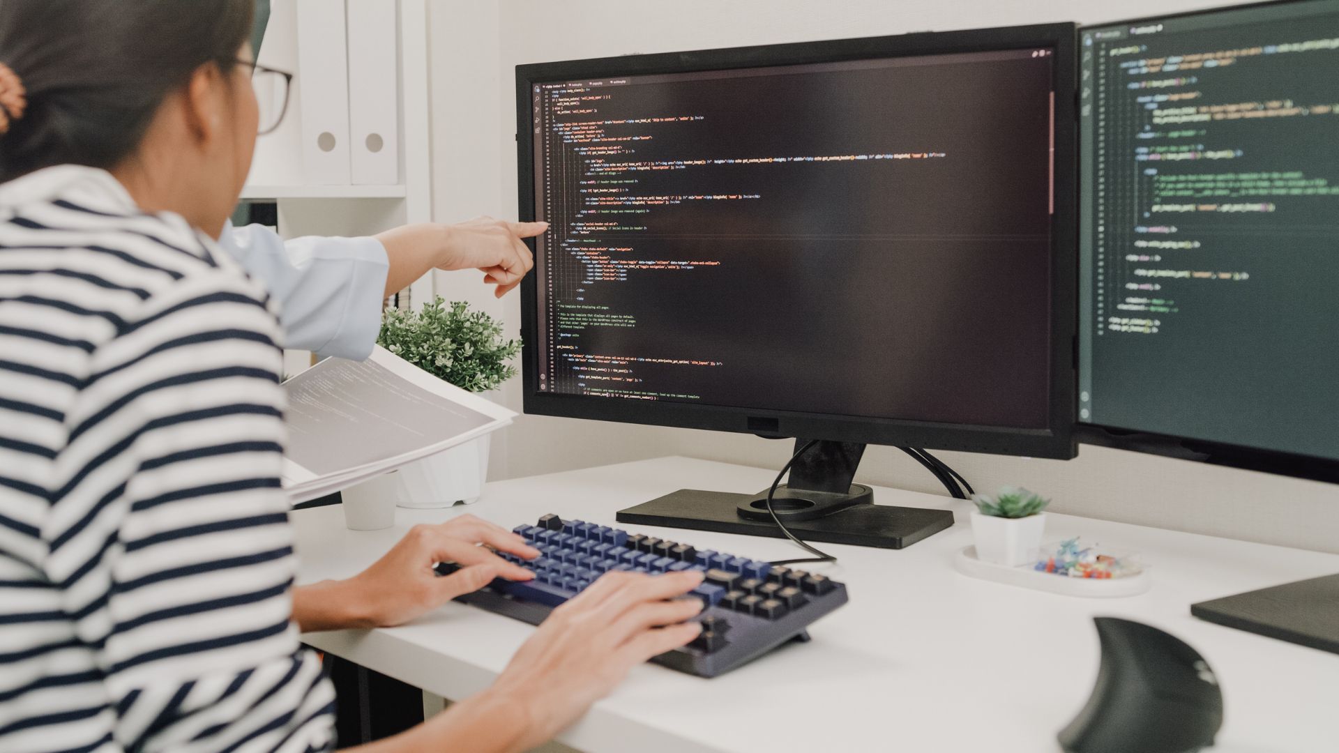 A woman is learning to code. A coworker we can't see is pointing at the woman's screen.