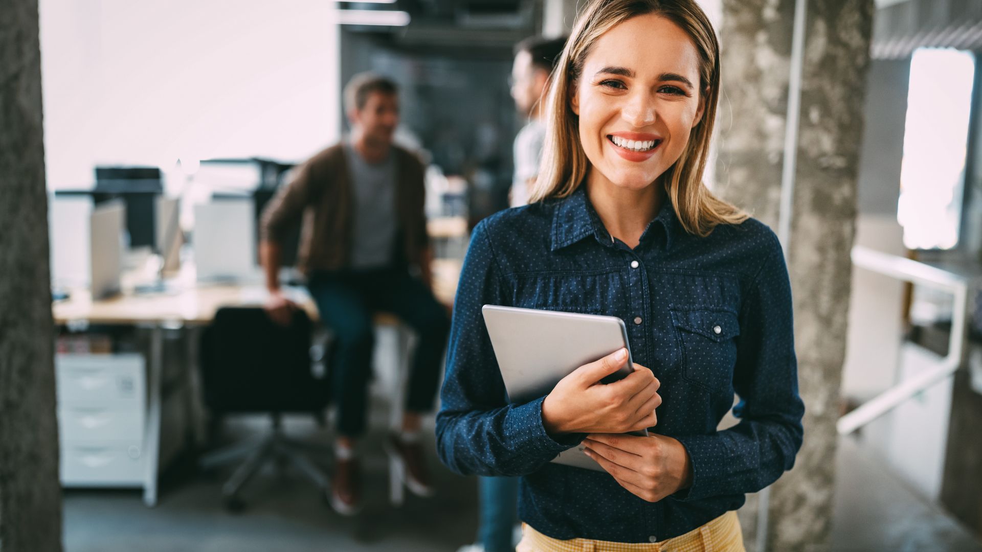 A smiling woman is holding a tablet, standing in an open office space.