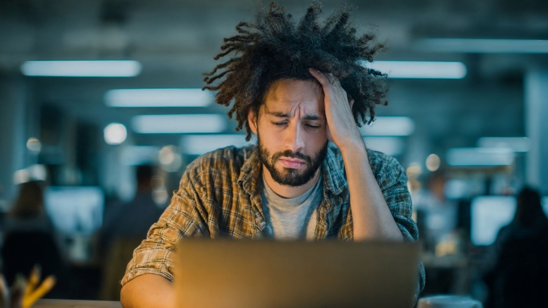 A manager is holding his head looking down at the laptop on his desk in the office.