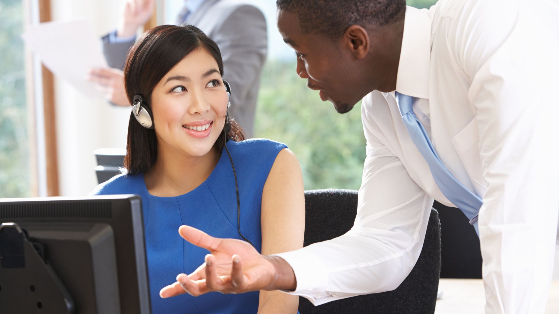 A manager is leaning over a seated employee having a conversation with them, showing them how to do something.