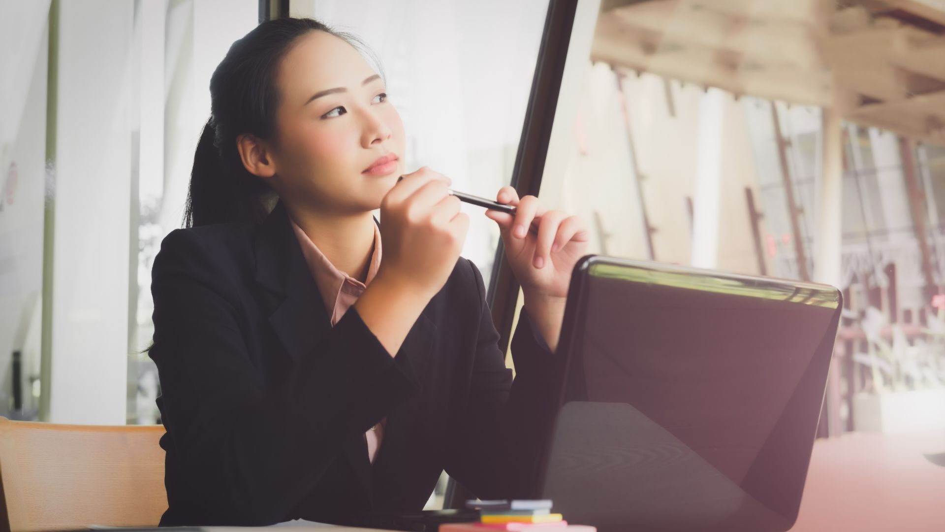 A woman is sitting in front of her laptop with a pencil in her hands, staring off - deep in thought.