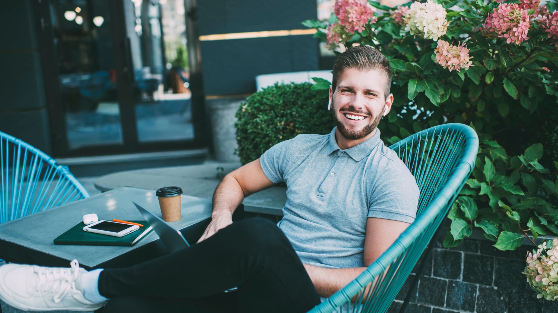 A man is sitting with his laptop in his lap outside at a table near a flower bush. He is smiling at the camera.