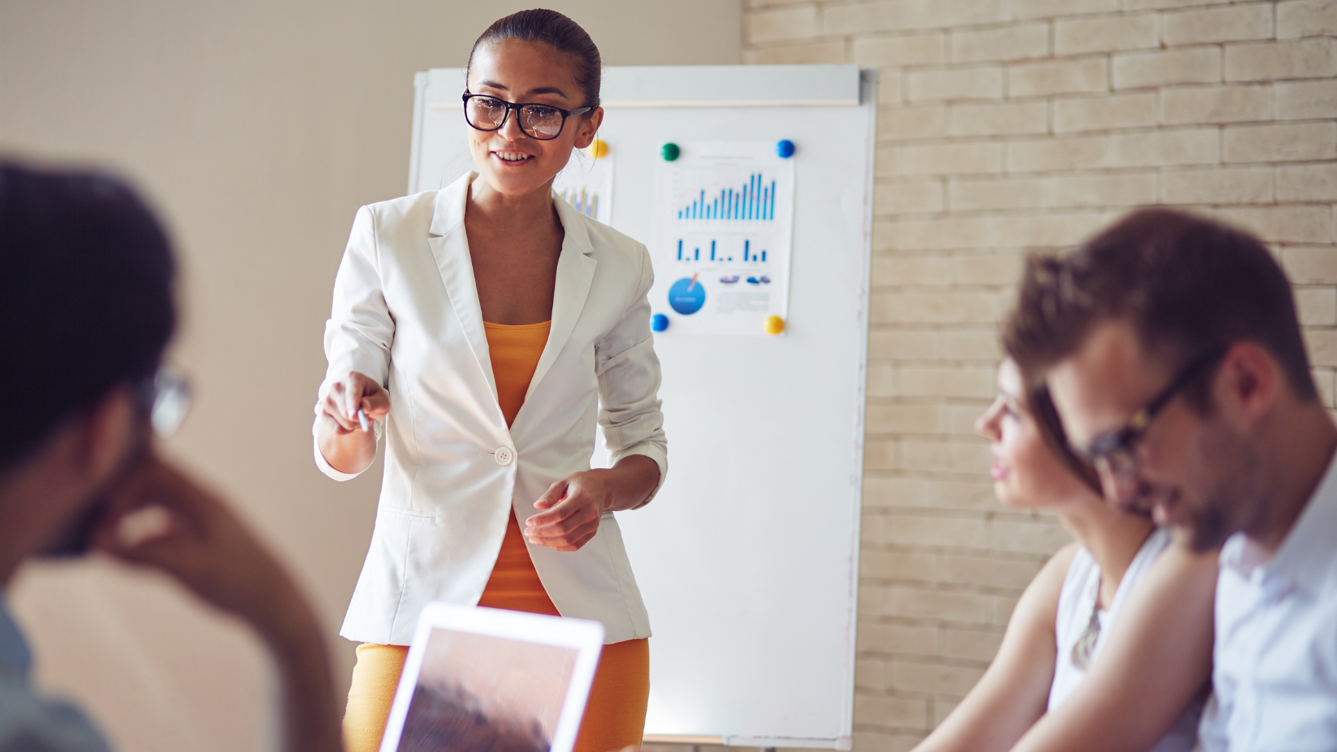 There's a woman standing in front of two people who are sitting down and she is presenting a chart that sits behind her.
