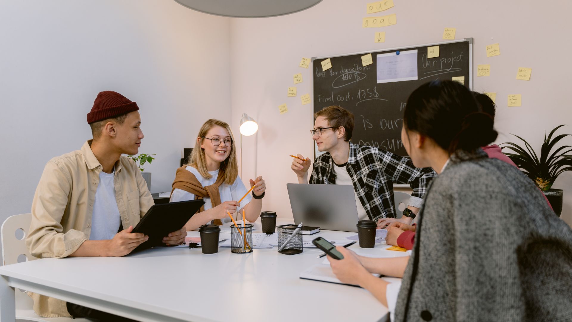 A diverse group of four young office workers are sitting around a conference table talking. There is a whiteboard and post it notes behind them.