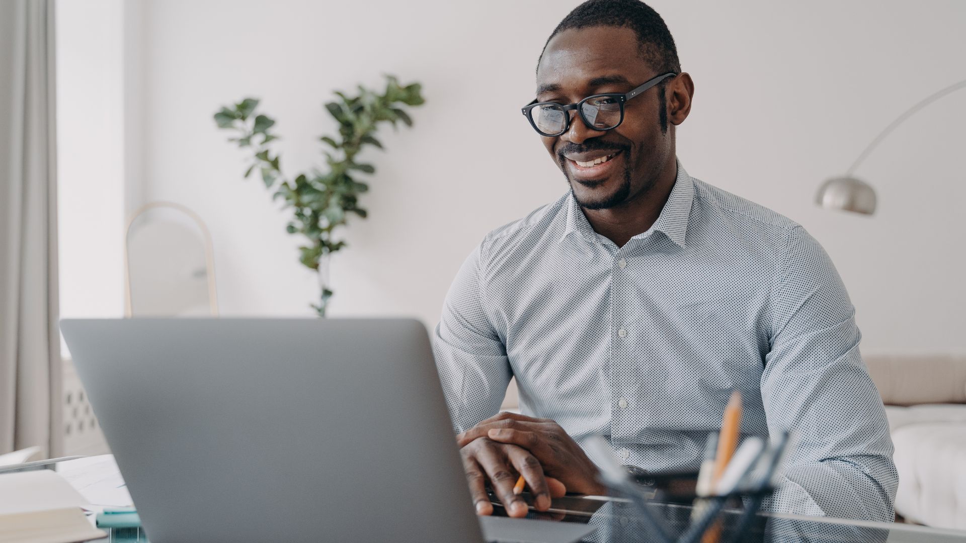 A young man wearing glasses is smiling while looking down at the live manager training on his laptop.