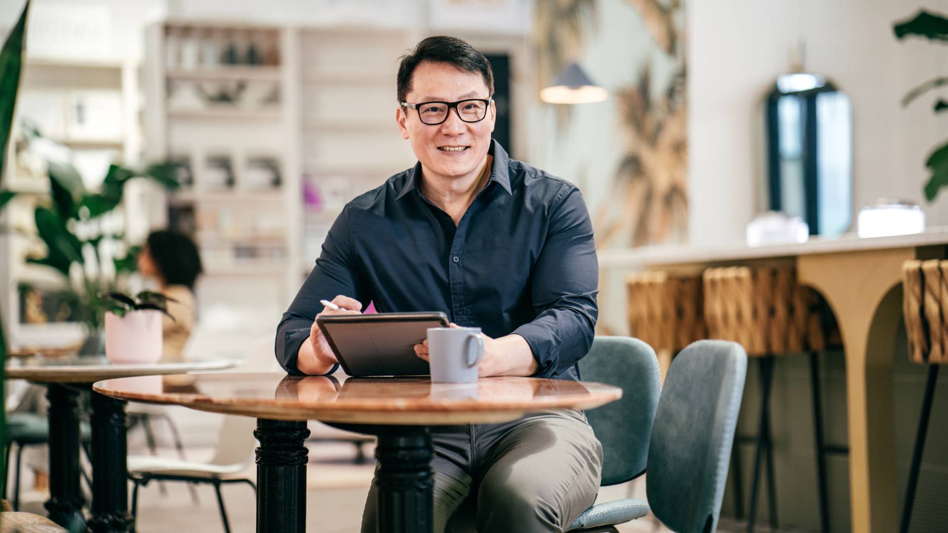 An Asian man with glasses is smiling at the camera while sitting at a cafe table holding a tablet.