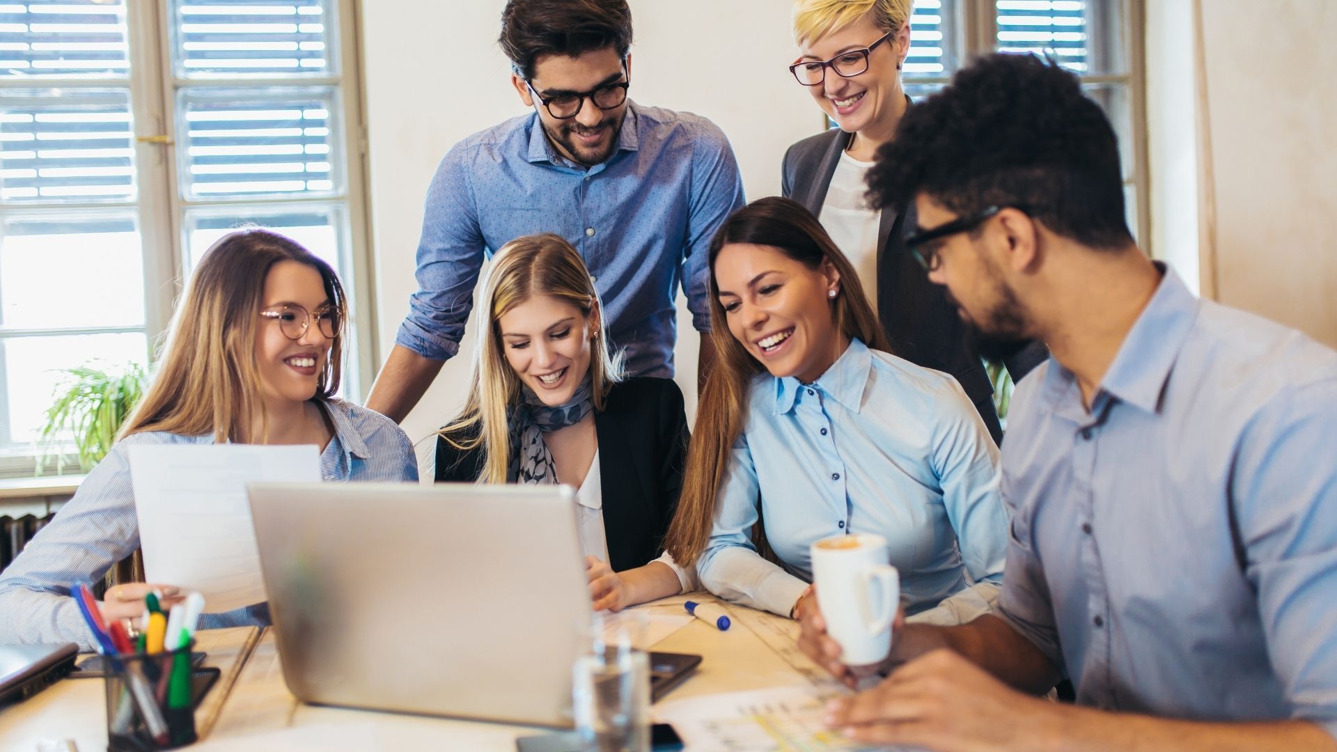 A diverse team of people are smiling and laughing, looking at the same laptop.