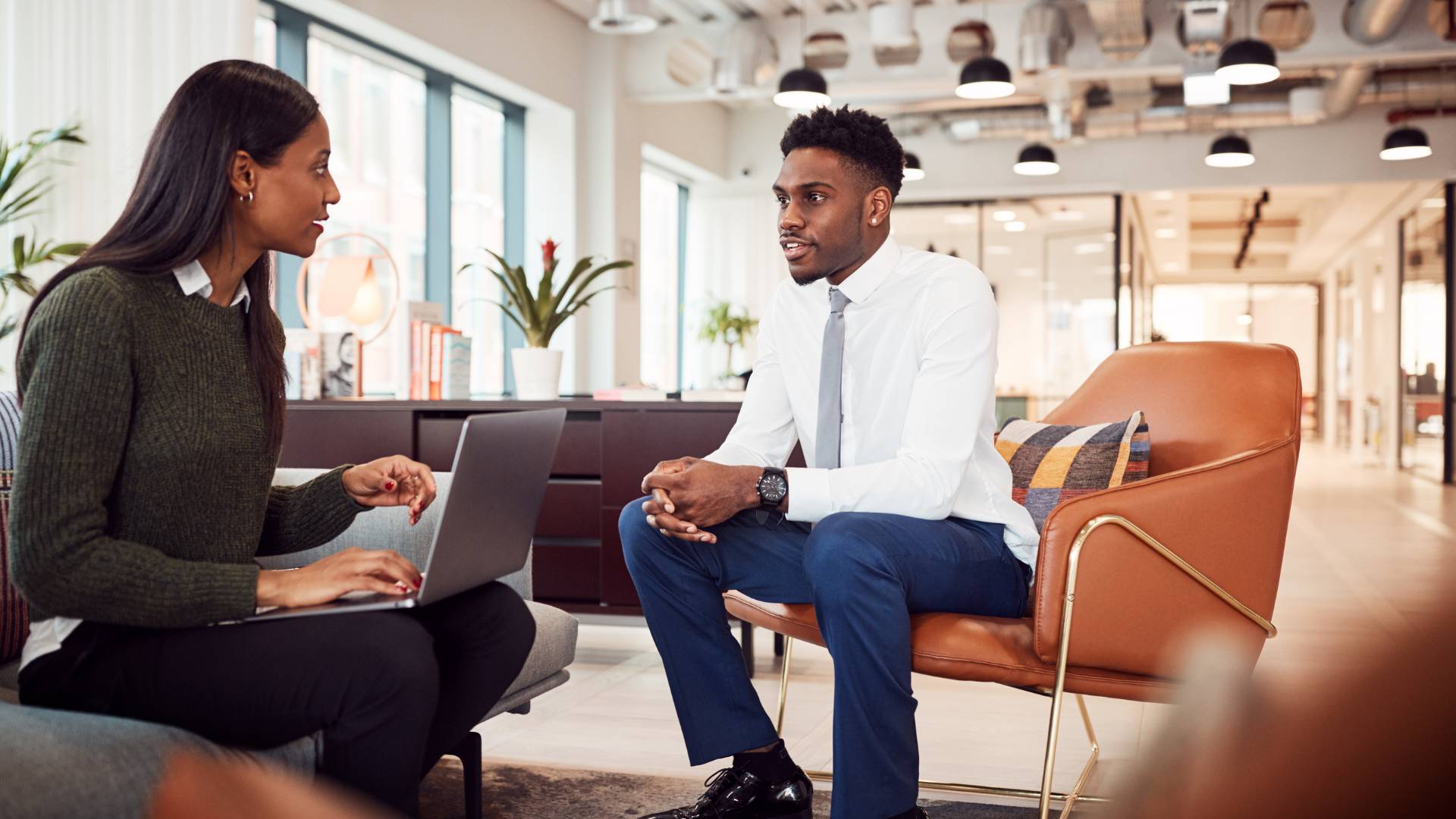 Two young professionals are sitting in a common area having a conversation. The woman on the left has a laptop on her left as she interviews the man sitting on the right.
