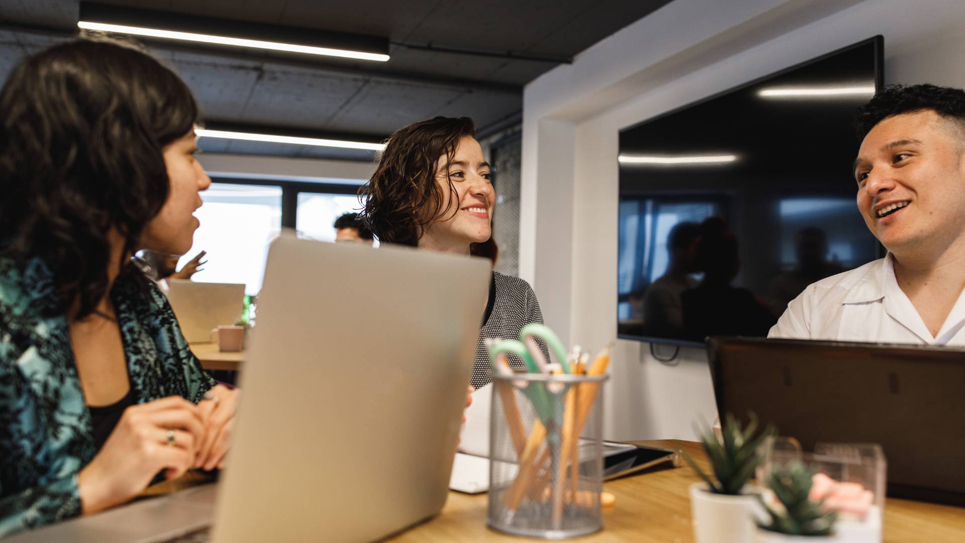 A group of 3 employees are working together in an office and smiling at each other over their laptops.