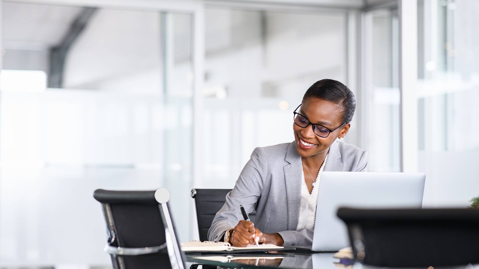 A woman is writing something in her notebook while sitting at a conference table with her laptop.