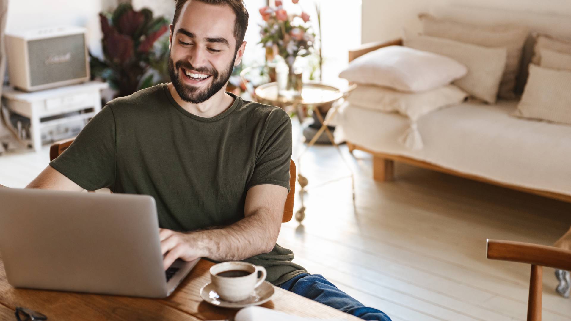 A young man is working on his laptop from a desk in his living room and is smiling.