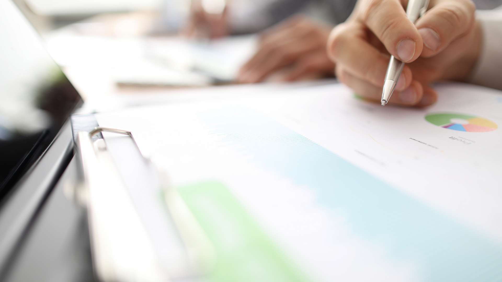 A close-up image of a hand holding a pen over a clipboard. The clipboard has data and graphs on it.