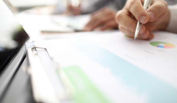 A close-up image of a hand holding a pen over a clipboard. The clipboard has data and graphs on it.