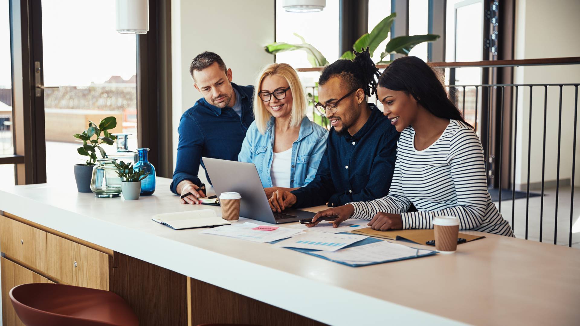 A group of 4 diverse employees are sitting on one side of a table and looking at a coworker's laptop.