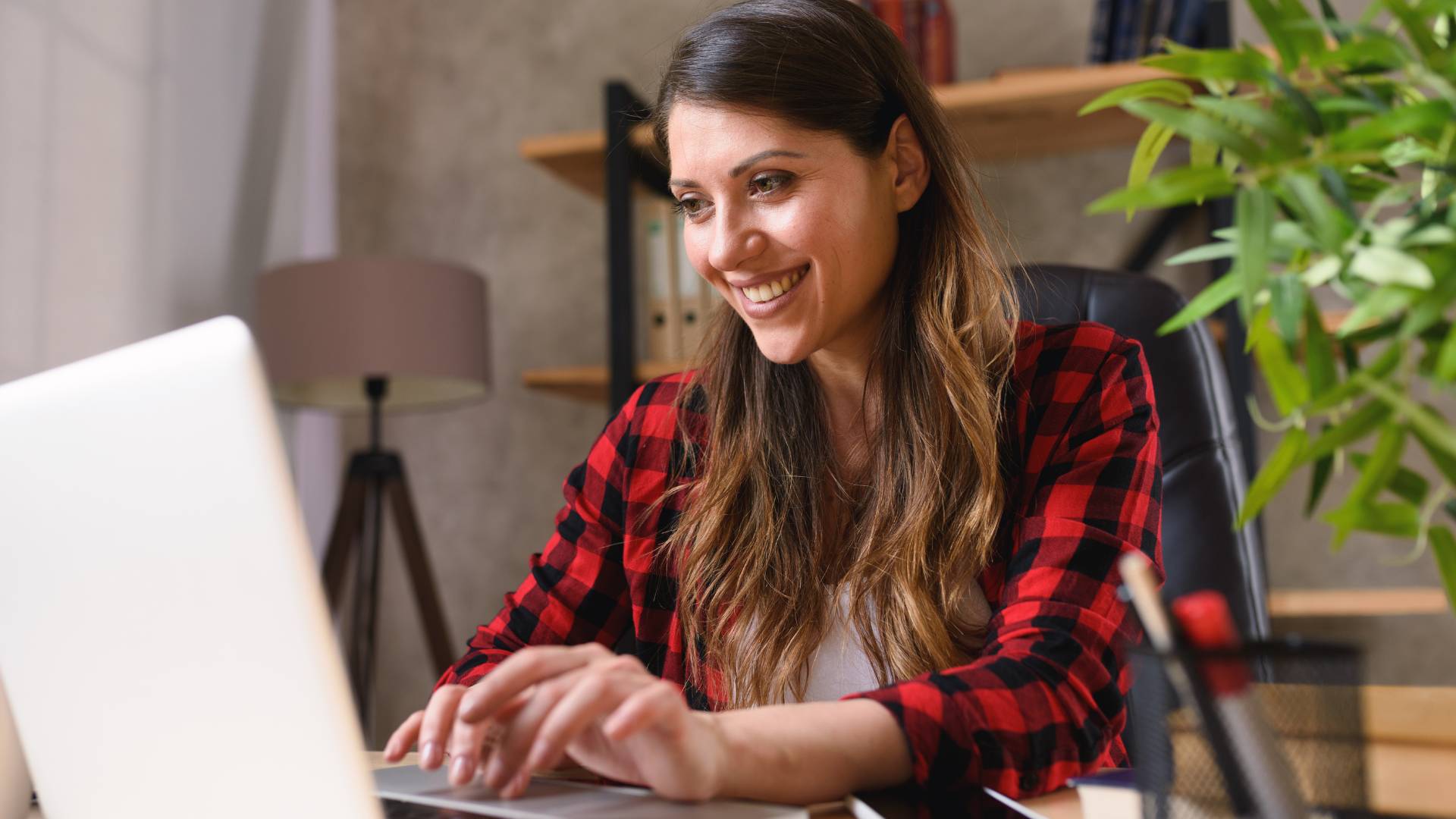 A woman is working from her home office and smiling at her laptop.
