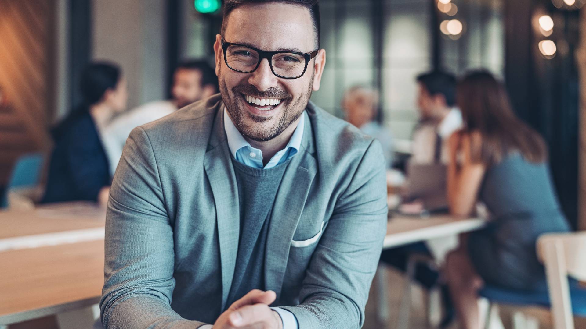 A man with glasses is smiling at the camera. Behind him coworkers are talking at a table.