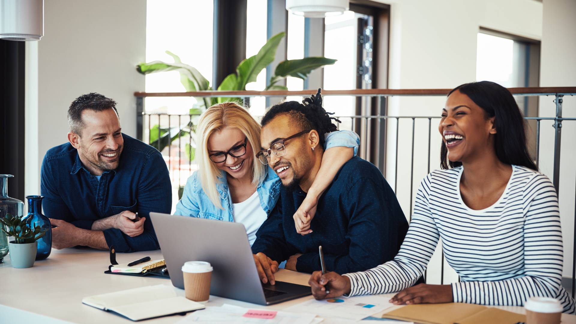 A group of employees in an office sitting around a table at a laptop, laughing and smiling.