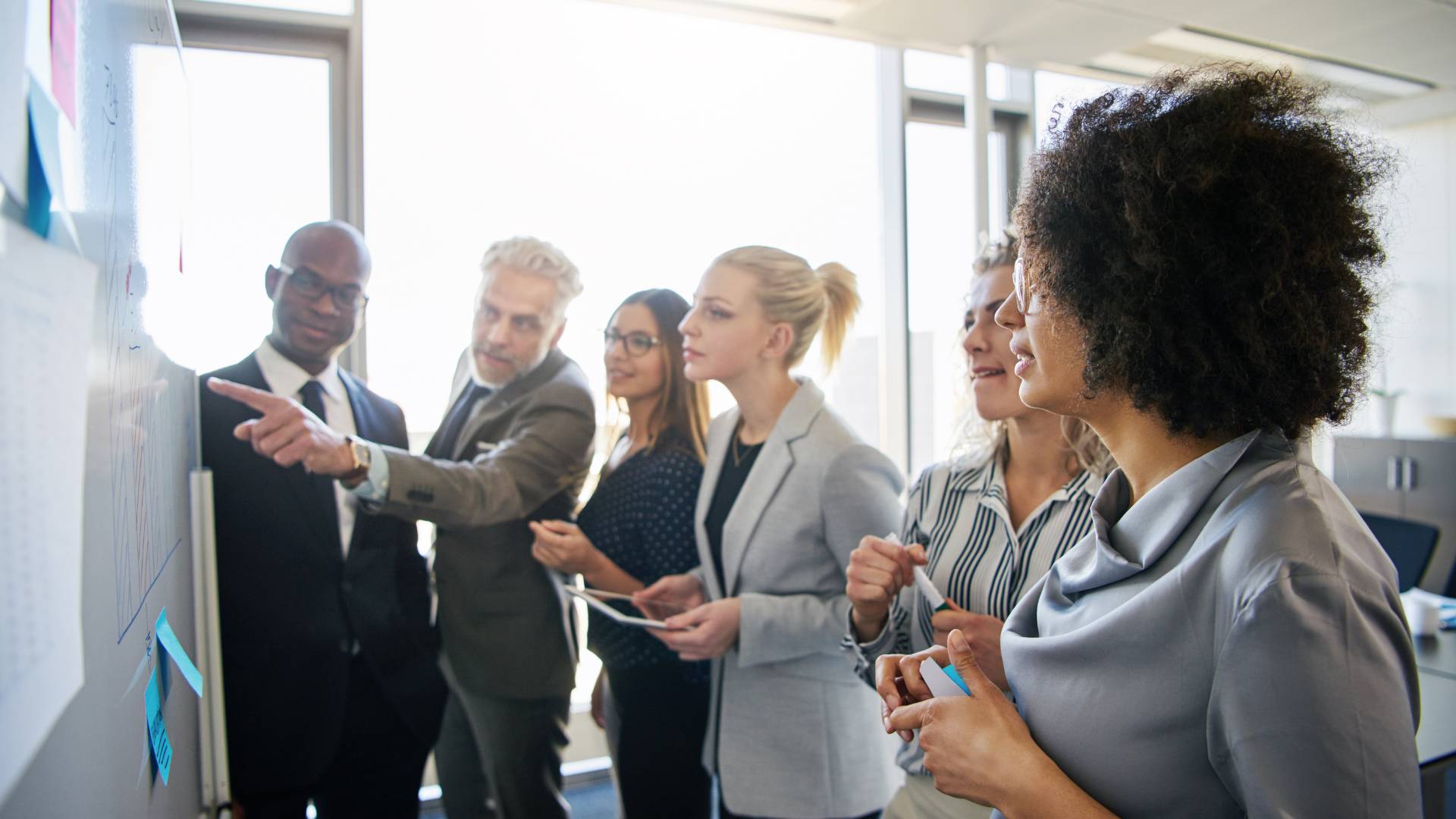 A diverse group of people is working around a white board. One man is pointing it at it.