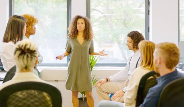 A Black woman is in a conference room standing up and conducting a meeting. There are six other people in the meeting.