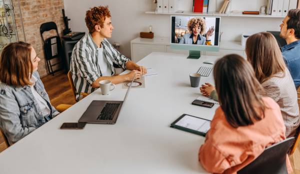 A team of people are seated around a conference table looking at a colleague on-screen at the end of the table.