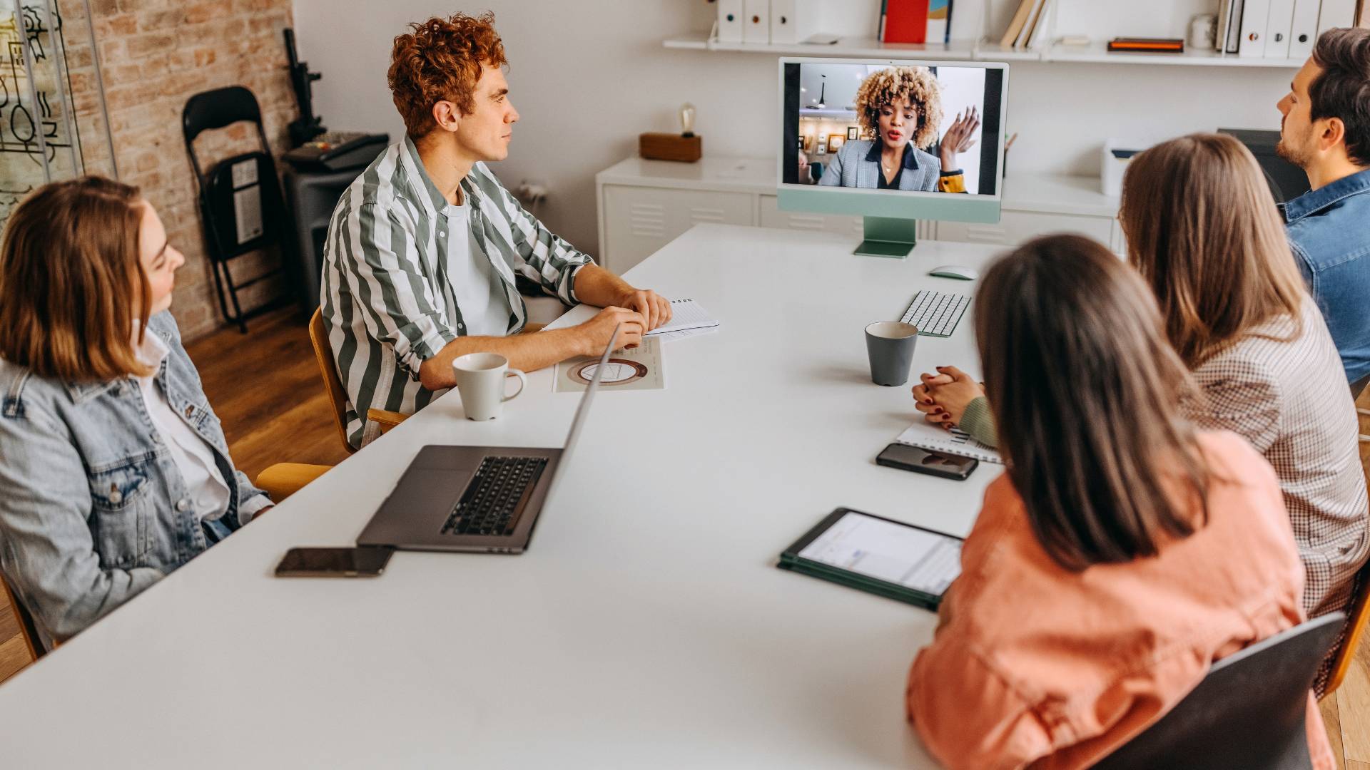 A team of people are seated around a conference table looking at a colleague on-screen at the end of the table.