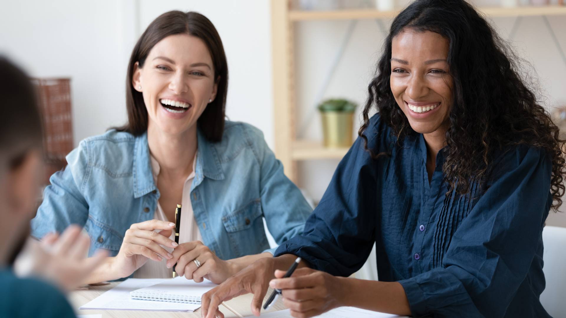 Two women are having a happy conversation over a conference table. We're looking over a mans shoulder at the two women smiling.