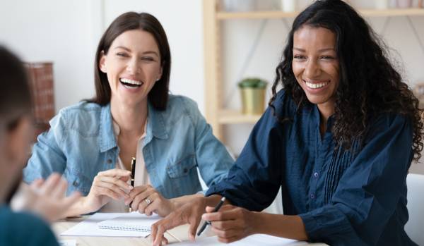 Two women are having a happy conversation over a conference table. We're looking over a mans shoulder at the two women smiling.