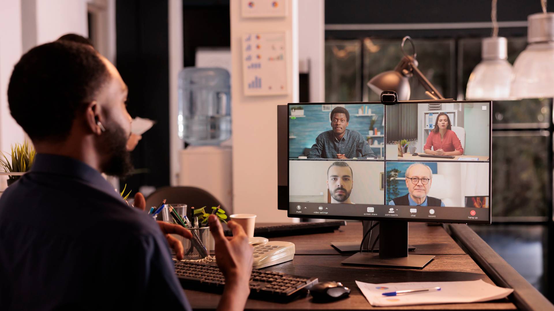 We are looking over a man's shoulder at his computer monitor on his desk while he talks to his team on video conference.