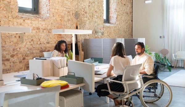 Three people sitting at a desk talking. One is in a wheelchair.