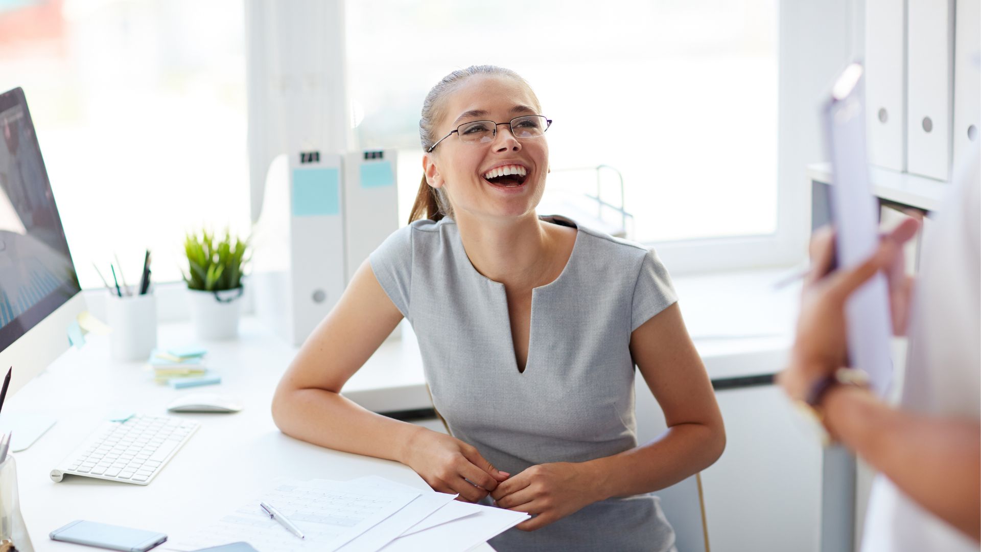 A joyful professional woman with glasses, wearing a grey sleeveless dress, is laughing while sitting at her desk in a brightly lit office. She is engaged in a lively conversation with a colleague, illustrating a positive workplace environment that supports employee mental health.