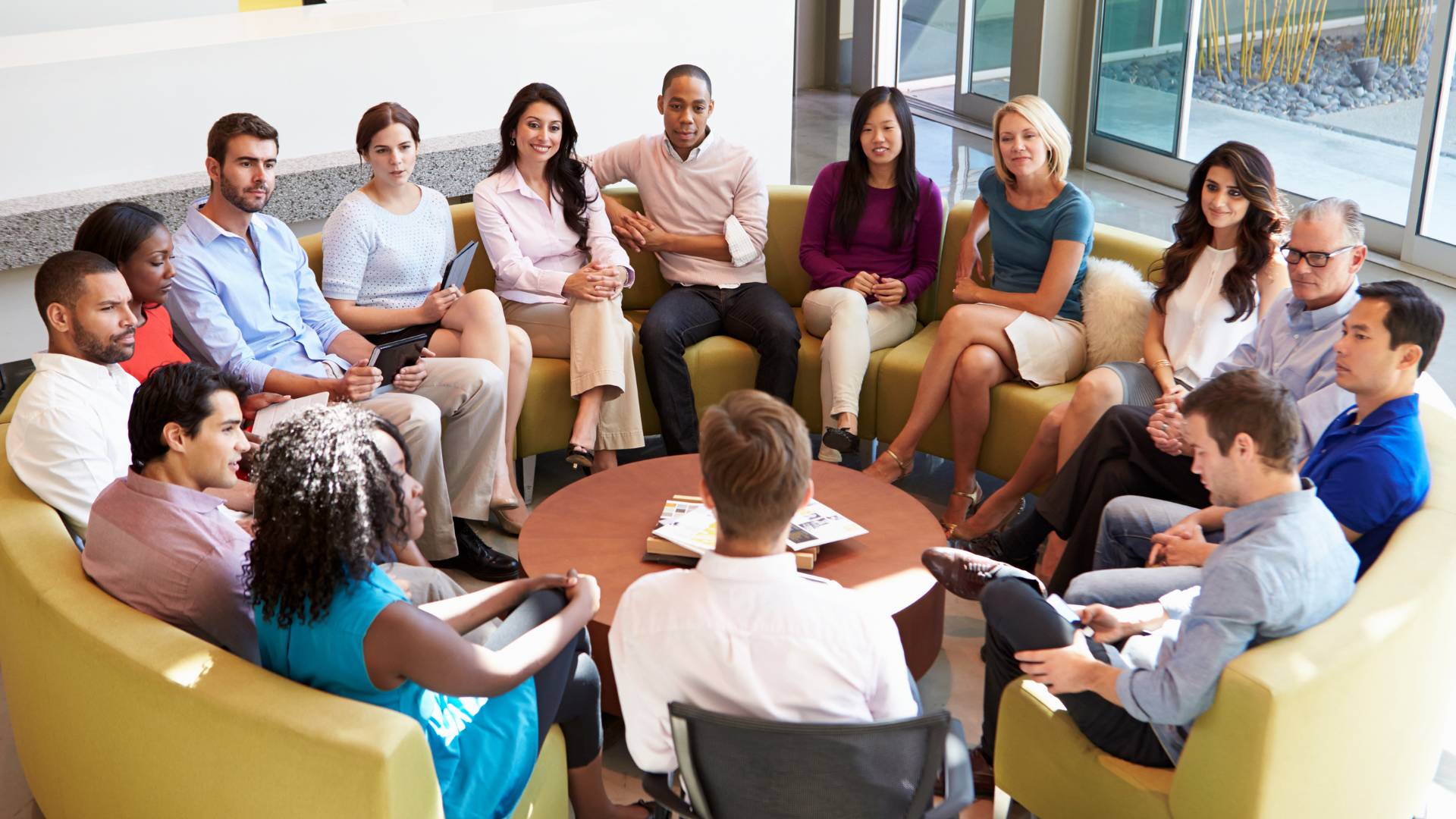 A diverse group of employees are sitting on a round couch in a common area during an employee development training.