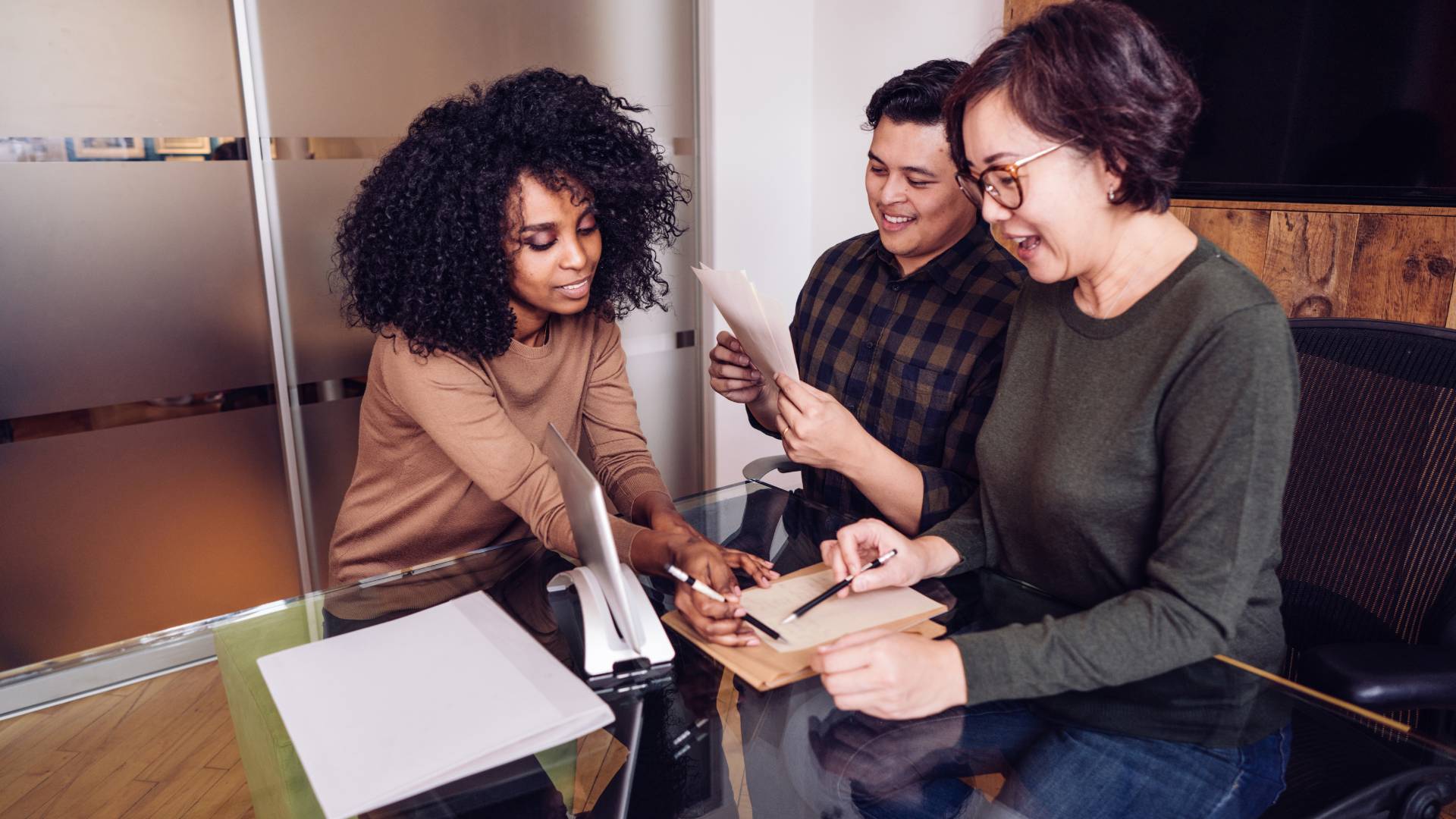 A group of 3 diverse employees are smiling and reviewing some paperwork together.