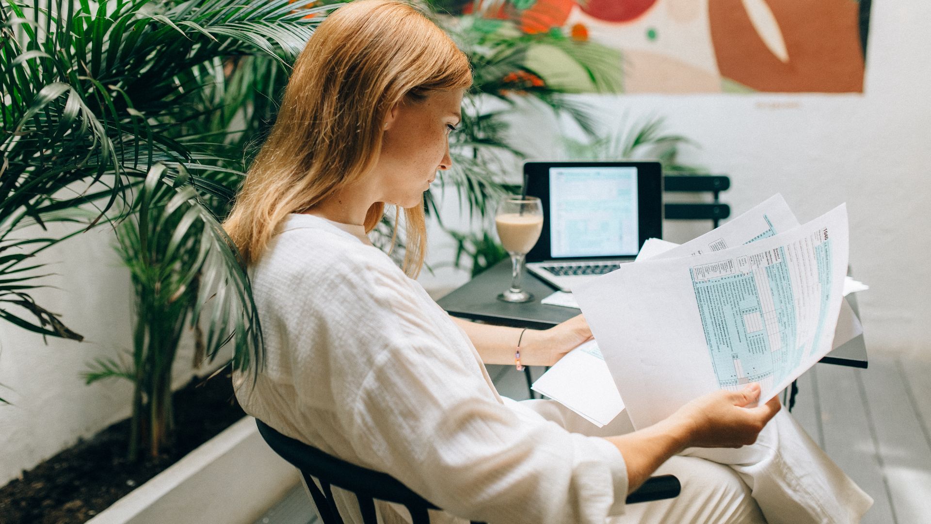 We are looking at the side profile of a woman reviewing a paper report. She is sitting next to a table with her laptop and a coffee.